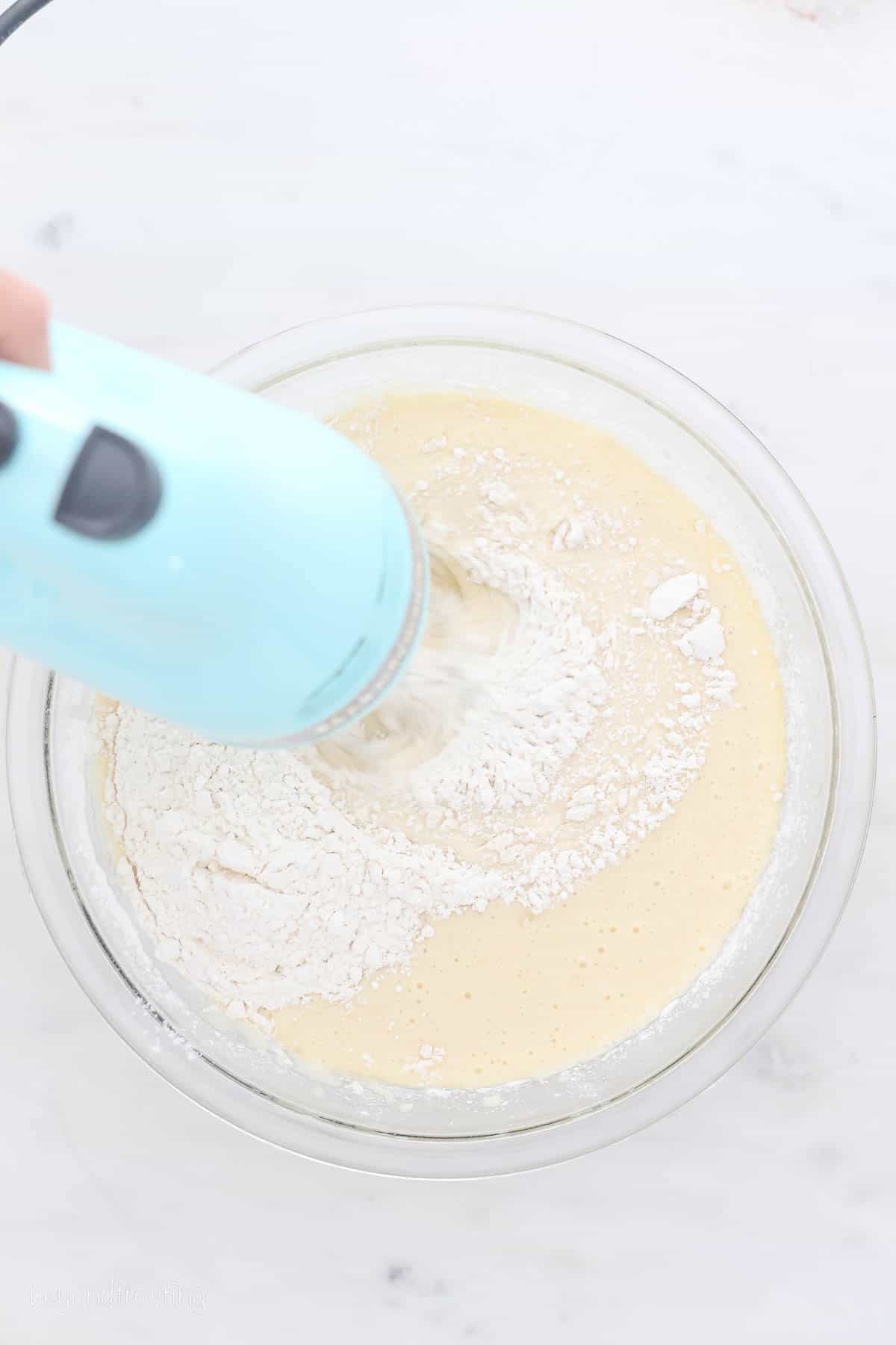 A hand blender is used to combine vanilla cake batter ingredients in a glass mixing bowl.