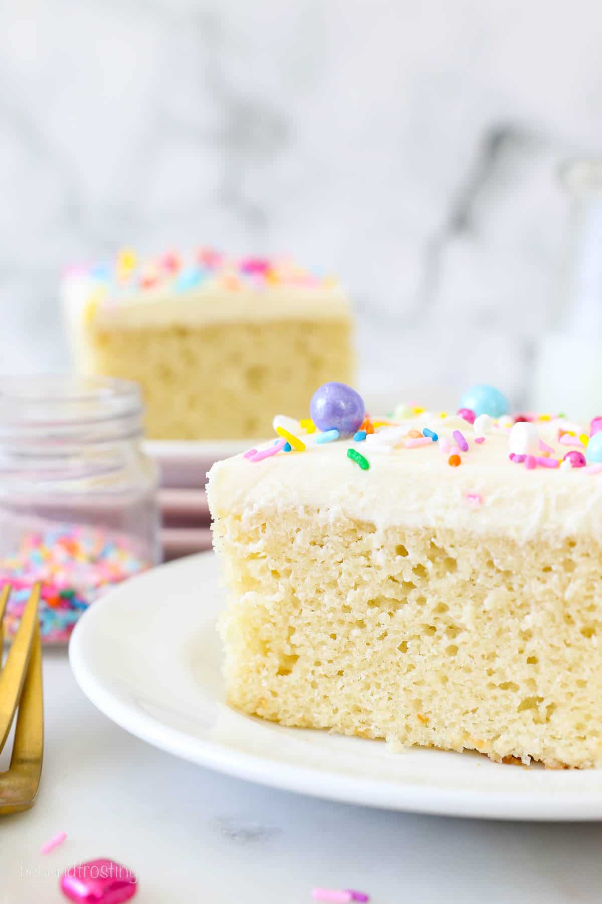 A slice of frosted vanilla cake on a white plate, garnished with rainbow sprinkles, with another slice of cake in the background.