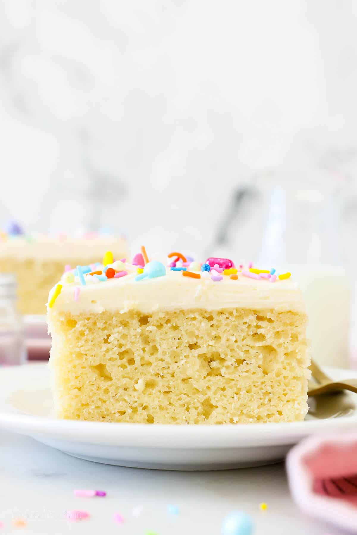 A slice of frosted vanilla cake on a white plate, garnished with rainbow sprinkles.