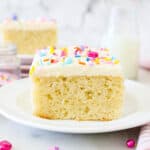 A slice of frosted vanilla cake on a white plate, garnished with rainbow sprinkles, with another slice of cake in the background.