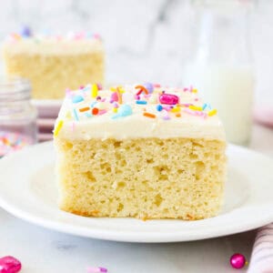 A slice of frosted vanilla cake on a white plate, garnished with rainbow sprinkles, with another slice of cake in the background.