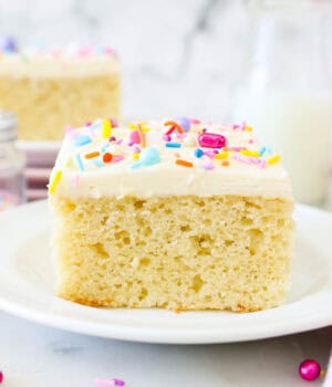 A slice of frosted vanilla cake on a white plate, garnished with rainbow sprinkles, with another slice of cake in the background.
