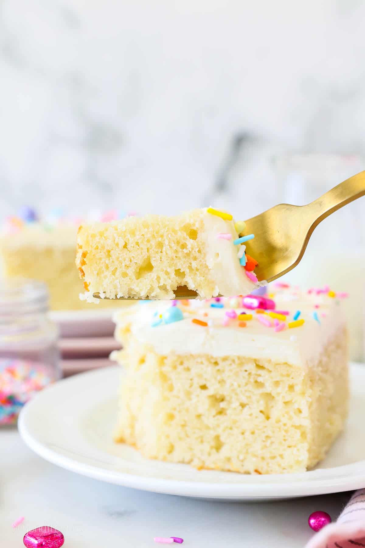 A forkful of vanilla cake held up in front of a slice of frosted cake garnished with rainbow sprinkles.