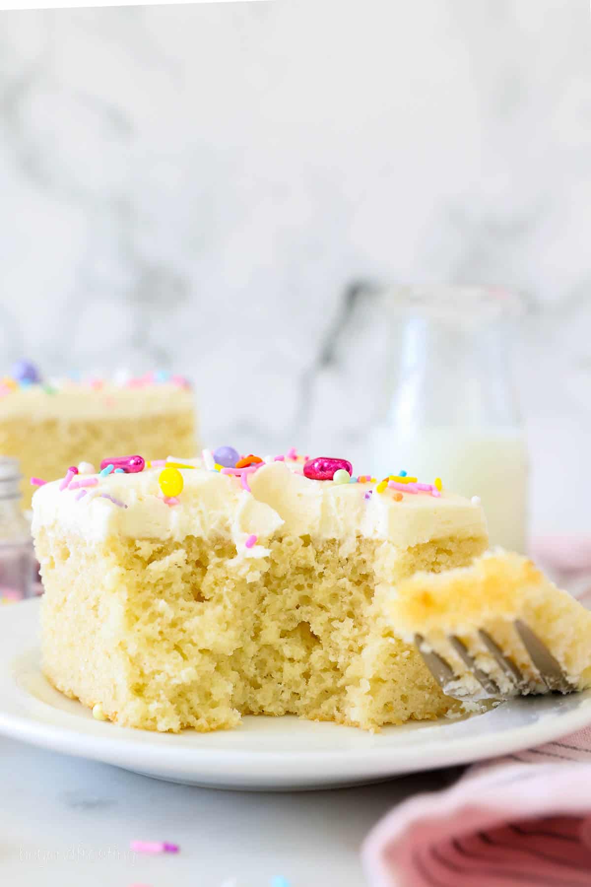 A slice of frosted vanilla cake topped with rainbow sprinkles on a white plate next to a forkful of cake.