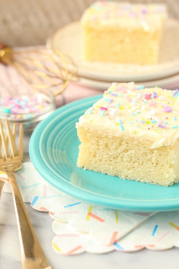 An overhead shot of a slice of vanilla cake on a light teal plate. The cake is garnished with colorful sprinkles and there's a couple gold forks in the background.