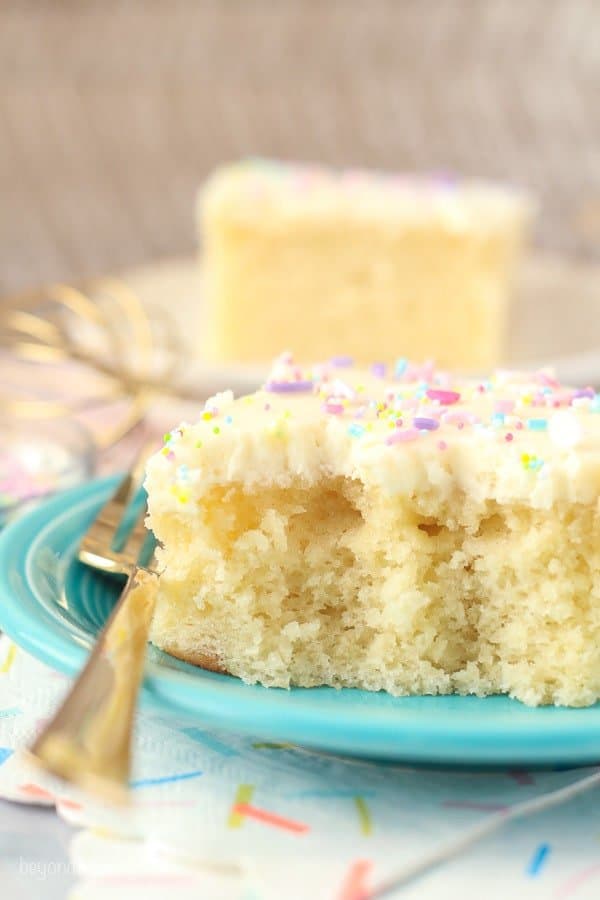 A gold fork sitting on a teal plate with a slice of vanilla cake. There's a few bites missing from the cake showing the spongy inside of the cake.