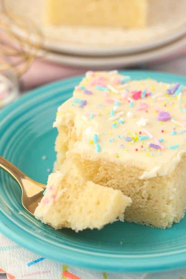 A slice of vanilla cake with frosting and sprinkles on a dark teal plate. The gold fork has a piece of cake on it, and there's a bite missing from the cake.