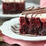 A close up shot of a double layer red velvet cake with a layer of brownie on the bottom and topped with whipped cream, drizzled with chocolate. The white plate is shitting on a red napkin