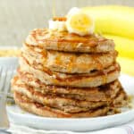 A stack of banana protein pancakes topped with banana slices and maple syrup on a white plate next to a fork.