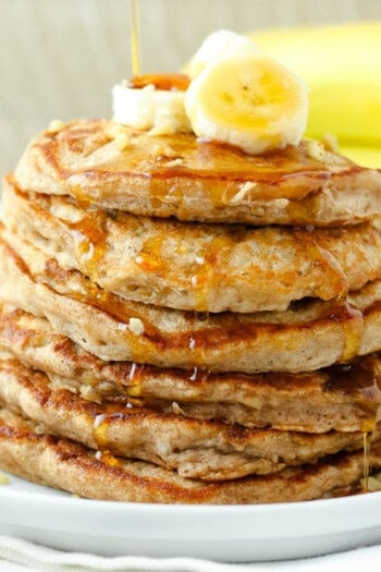 A stack of banana protein pancakes topped with banana slices and maple syrup on a white plate next to a fork.