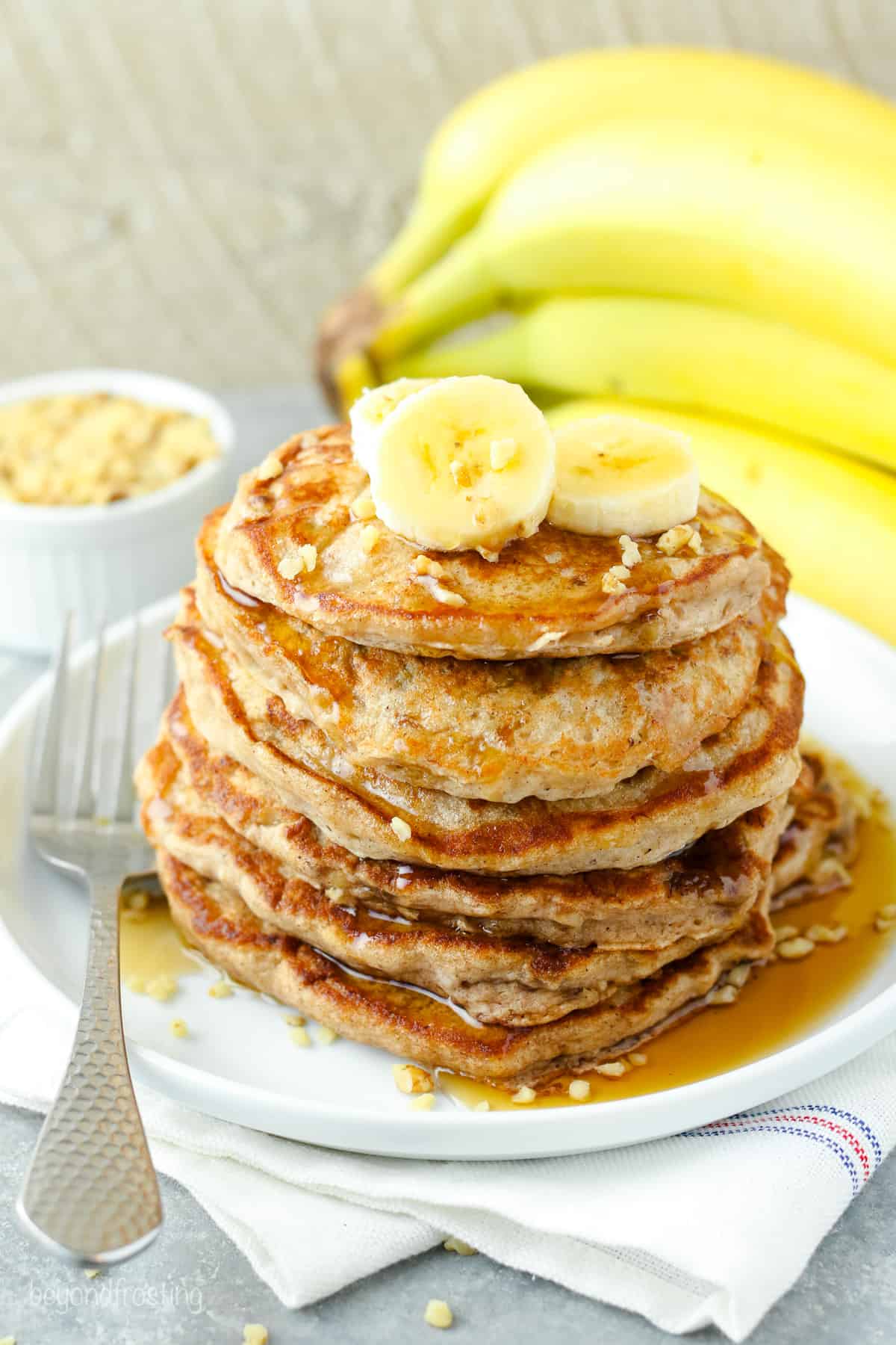 A stack of banana protein pancakes topped with banana slices and maple syrup on a white plate, with a bunch of bananas in the background.