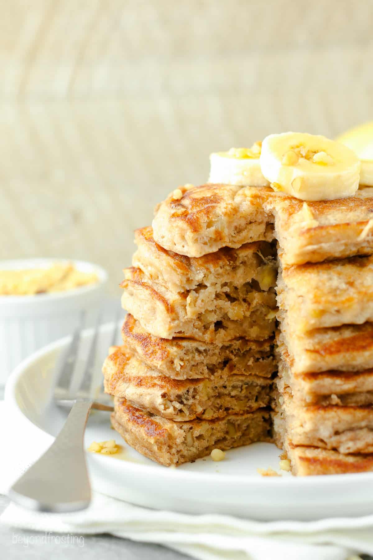A stack of banana protein pancakes topped with banana slices and maple syrup, with a large slice cut out, on a white plate next to a fork.