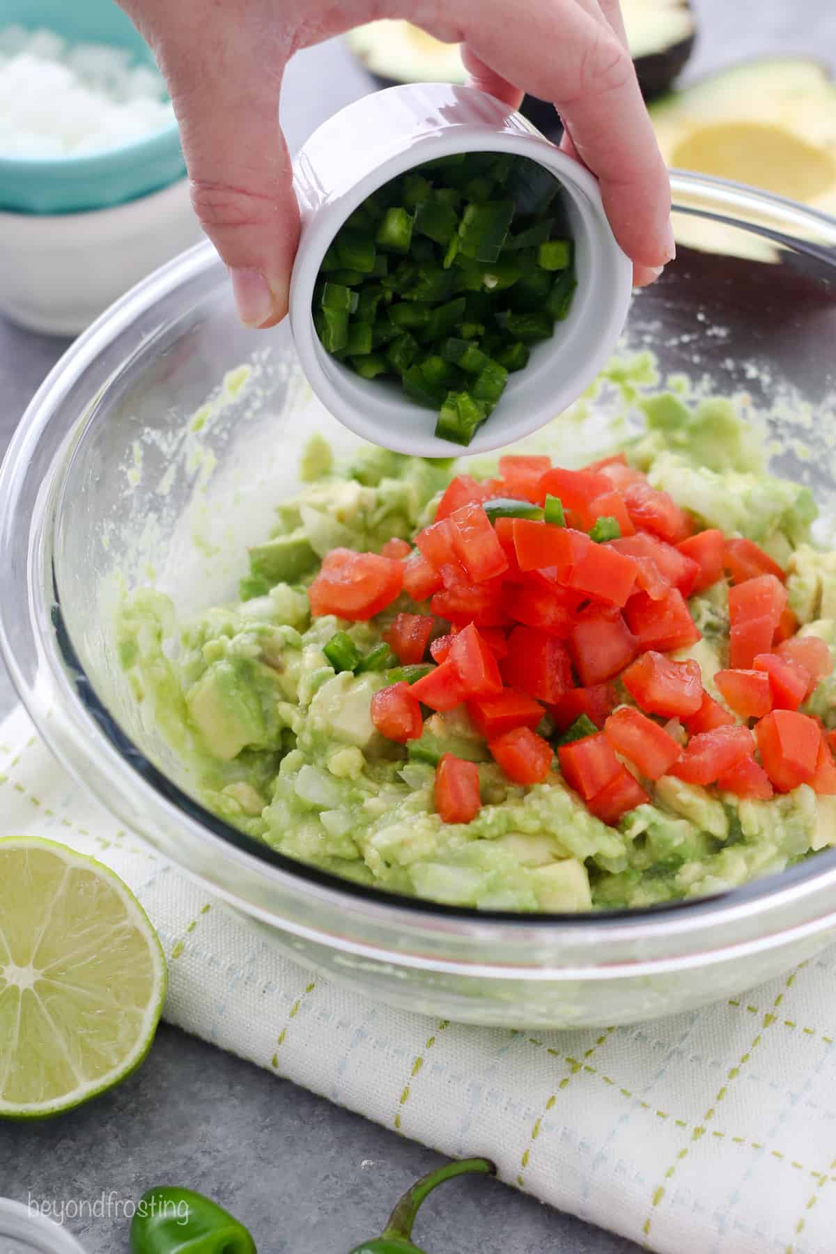 A hand dumping a small dish of diced jalapenos into a bowl with mashed avocado and diced tomatoes.