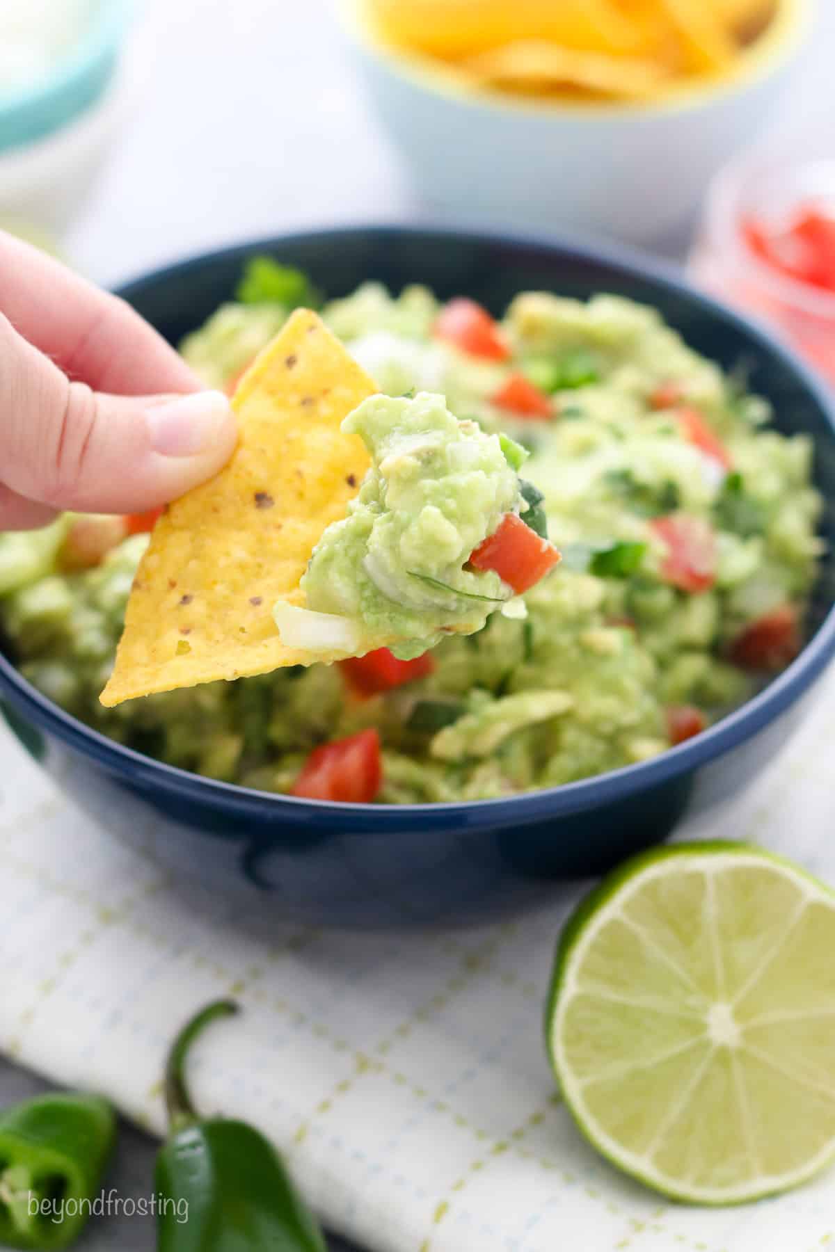 Close up of a hand holding a tortilla chip dipped in guacamole, with a bowl of guacamole in the background next to one half of a lime.