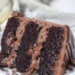 A close up shot of a 3 layer chocolate cake with chocolate frosting on a white rimmed plate