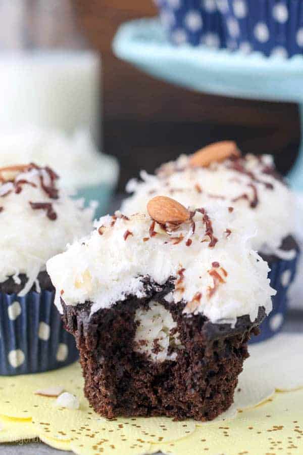 A chocolate cupcake with a bite taken out of it. The center of the cupcake shows a coconut filling and it has a coconut frosting garnished with an Almond