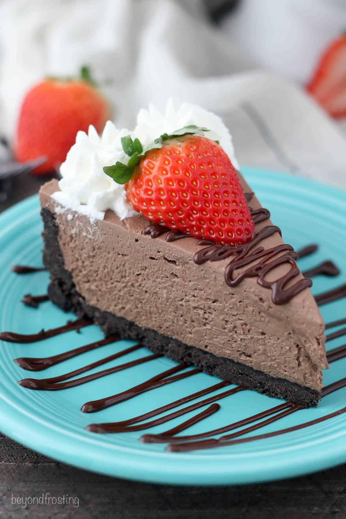 A slice of no-bake chocolate cheesecake topped with a strawberry and whipped cream, on a teal plate drizzled with chocolate sauce.