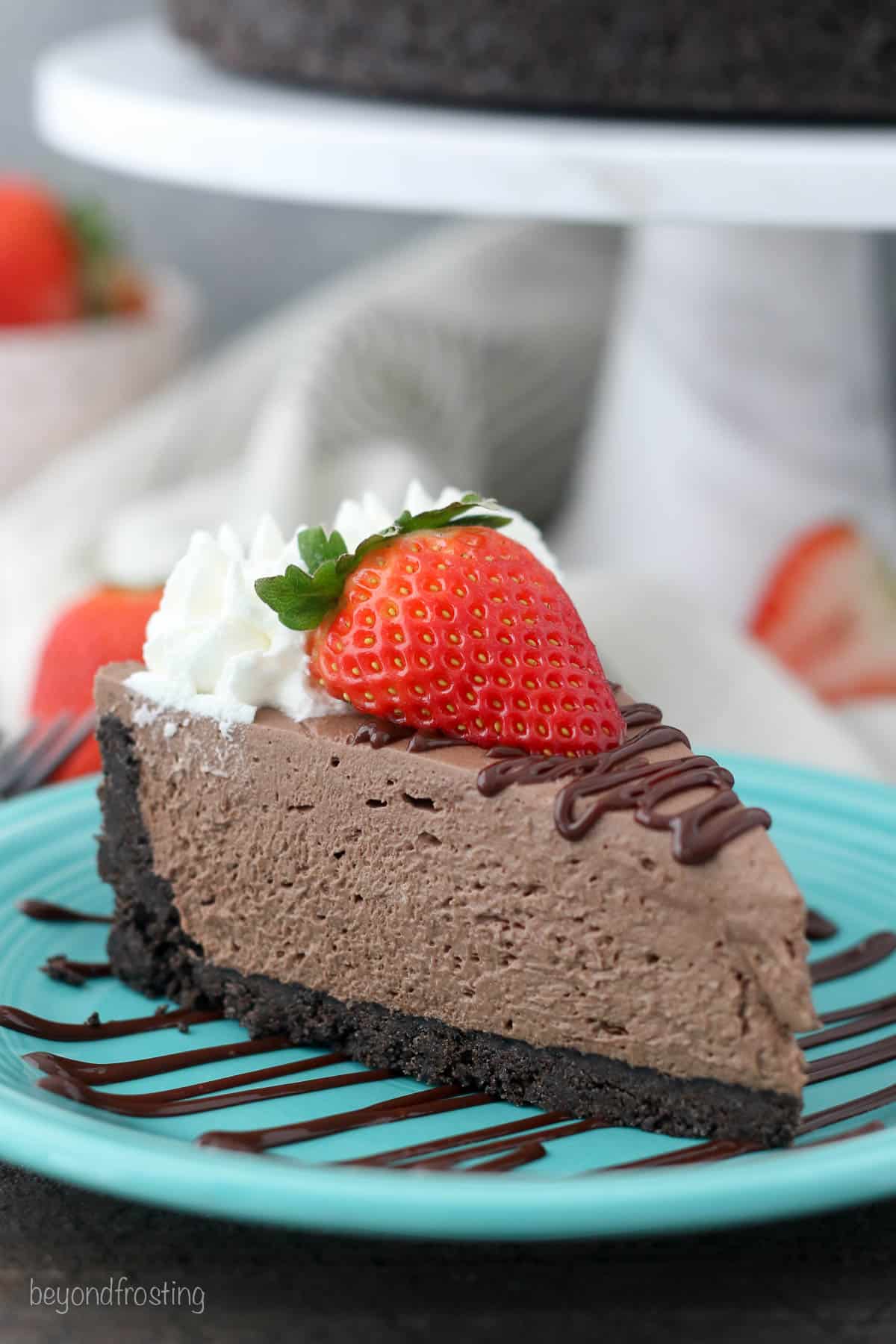 A slice of no-bake chocolate cheesecake topped with a strawberry and whipped cream, on a teal plate drizzled with chocolate sauce.