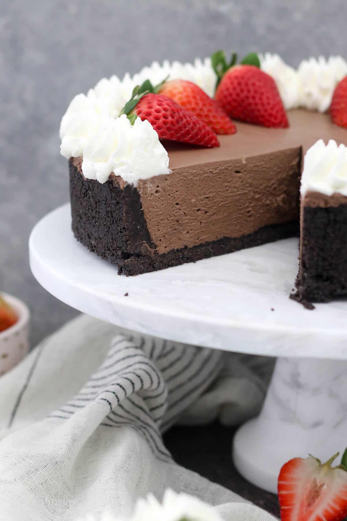 No-bake chocolate cheesecake topped with whipped cream and strawberries, with one slice missing on a cake stand.