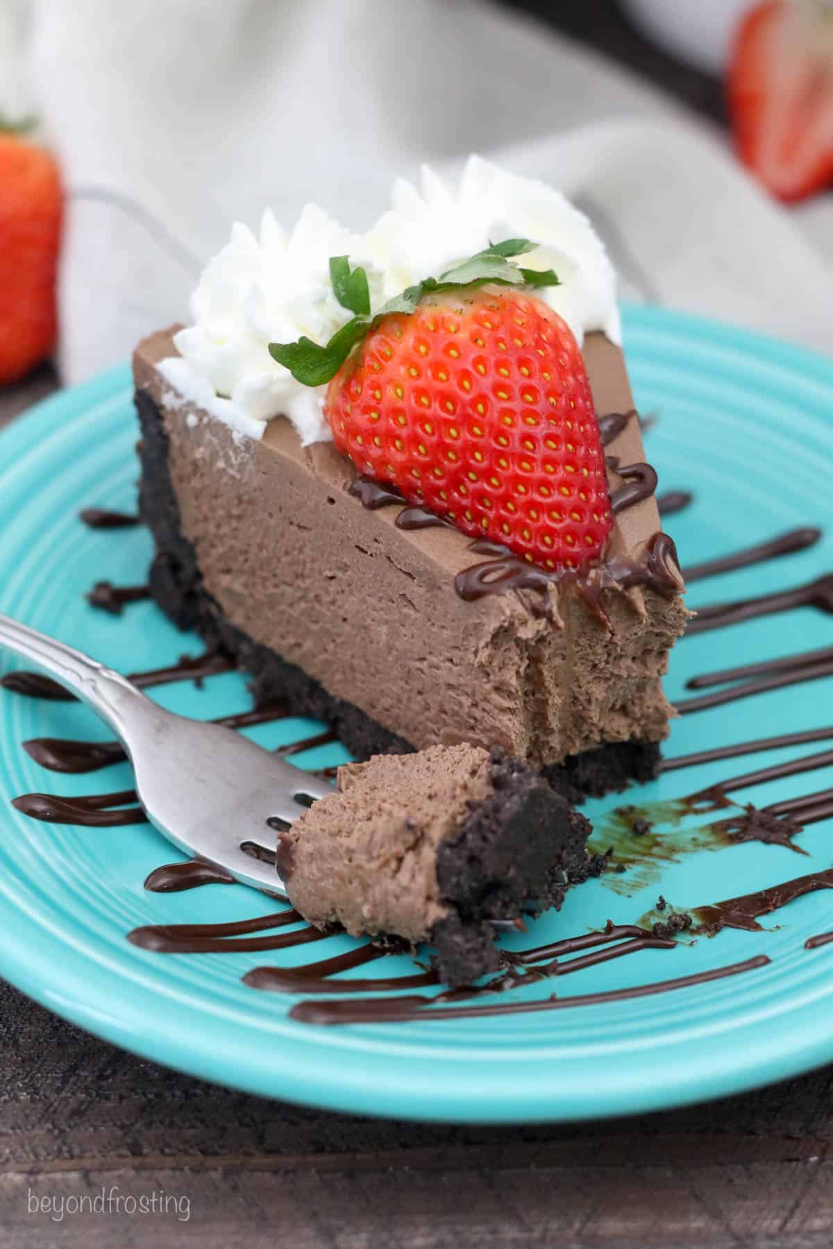 A slice of no-bake chocolate cheesecake on a teal plate topped with a strawberry and whipped cream, with a forkful missing.