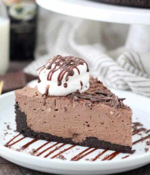 A slice of No-Bake Baileys Cheesecake on a white plate drizzled with chocolate and topped with whipped cream