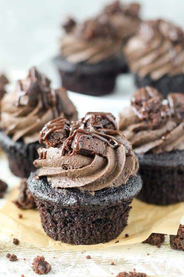 Three chocolate cupcakes topped with chocolate frosting, brownie pieces and hot fudge with three more cupcakes in the background