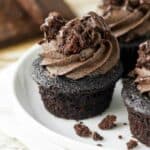 A chocolate cupcake on a white cake plate topped with brownie frosting and brownie chunks