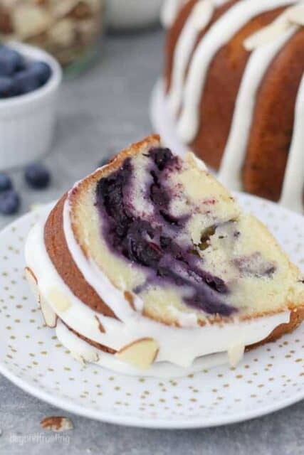 A slice of blueberry almond pound cake laying on a gold polka dot plate covered with a cream cheese glaze and sliced almonds