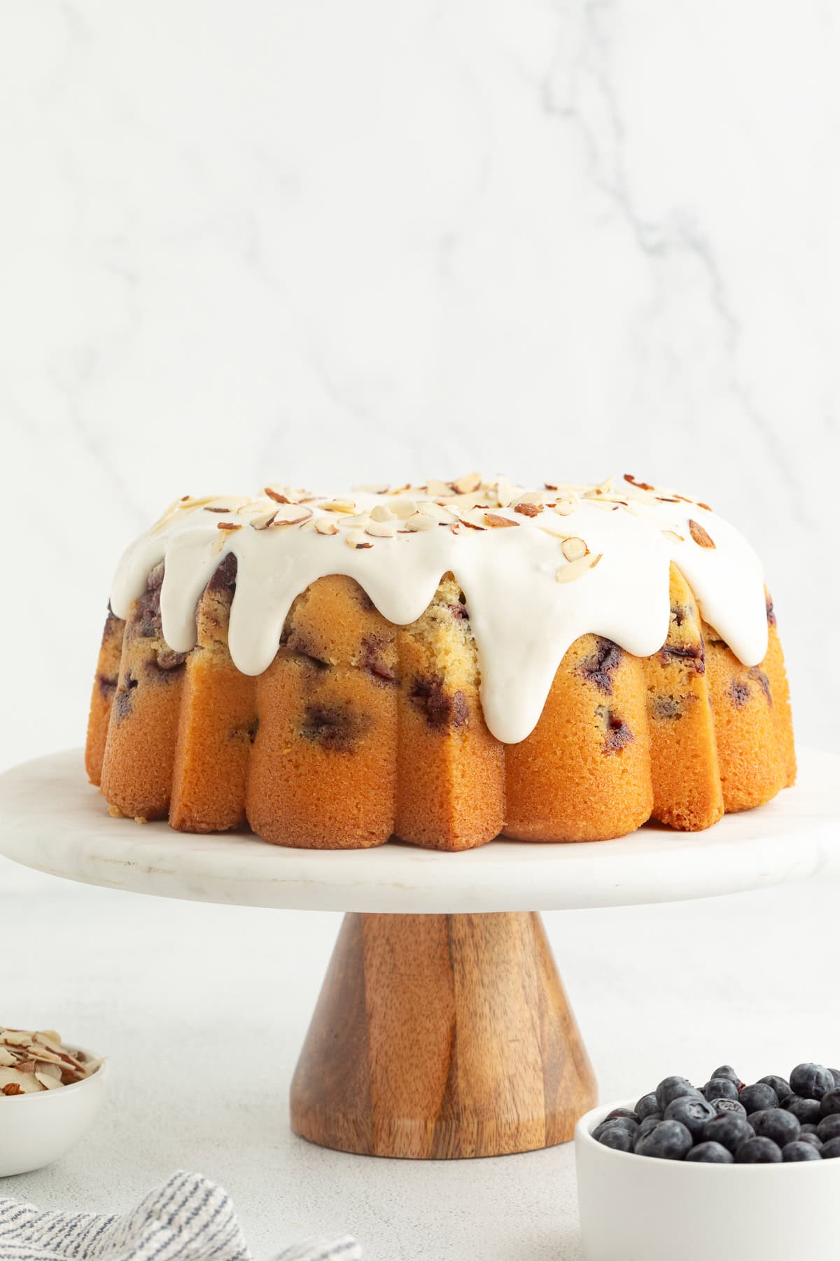 This blueberry bundt cake is a moist pound cake recipe with added almond flavor and juicy blueberries, topped with tangy cream cheese icing. Blueberry bundt cake topped with cream cheese icing and slivered almonds on a cake stand.