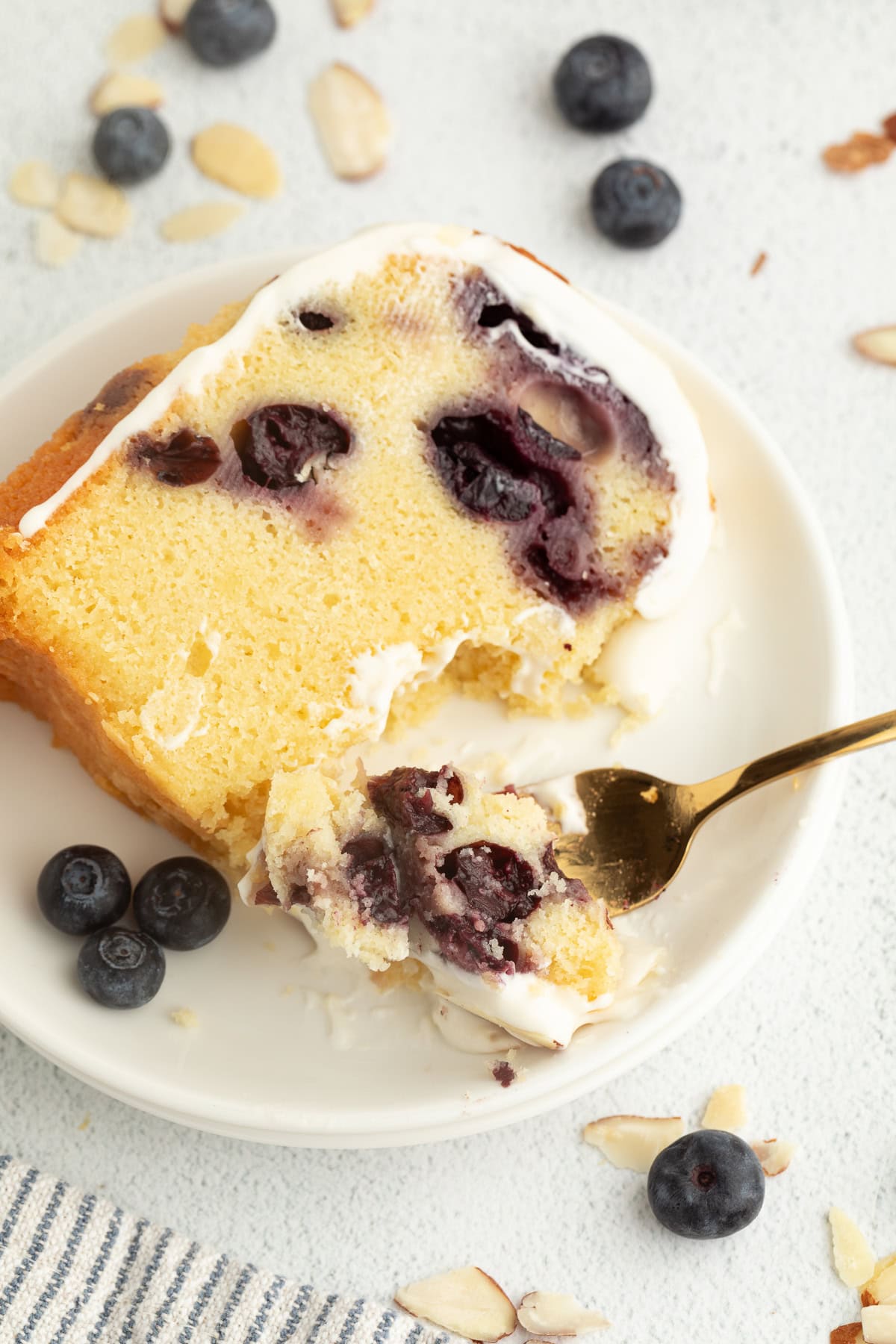 This blueberry bundt cake is a moist pound cake recipe with added almond flavor and juicy blueberries, topped with tangy cream cheese icing. Overhead view of a forkful of cake resting next to a slice of blueberry bundt cake on a plate.