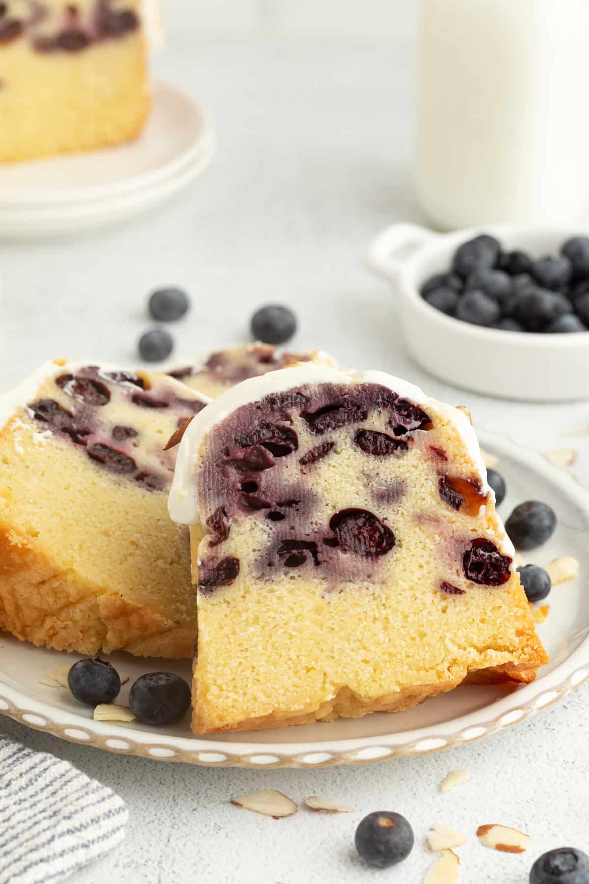 This blueberry bundt cake is a moist pound cake recipe with added almond flavor and juicy blueberries, topped with tangy cream cheese icing. Two slices of blueberry bundt cake with cream cheese icing on a plate.
