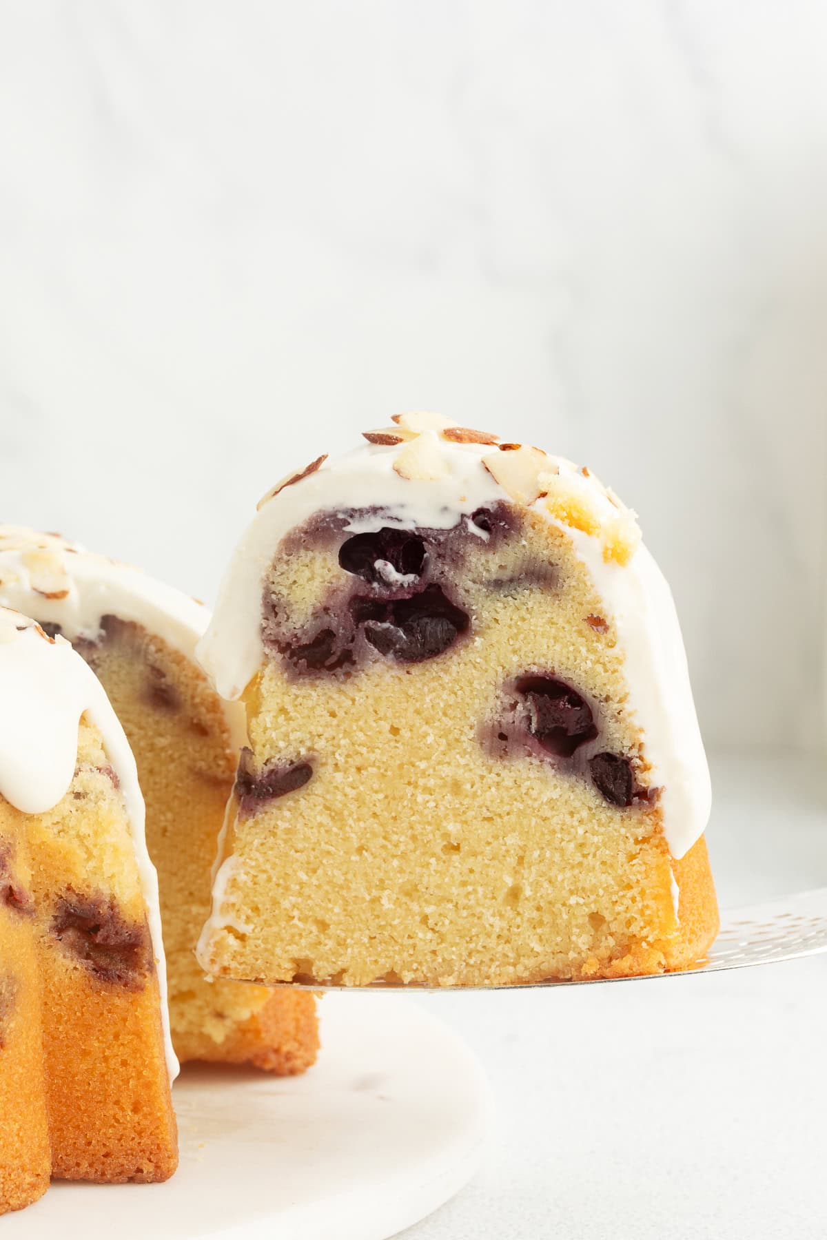 This blueberry bundt cake is a moist pound cake recipe with added almond flavor and juicy blueberries, topped with tangy cream cheese icing. A slice of blueberry almond bundt cake topped with cream cheese icing being lifted from the rest of the cake.