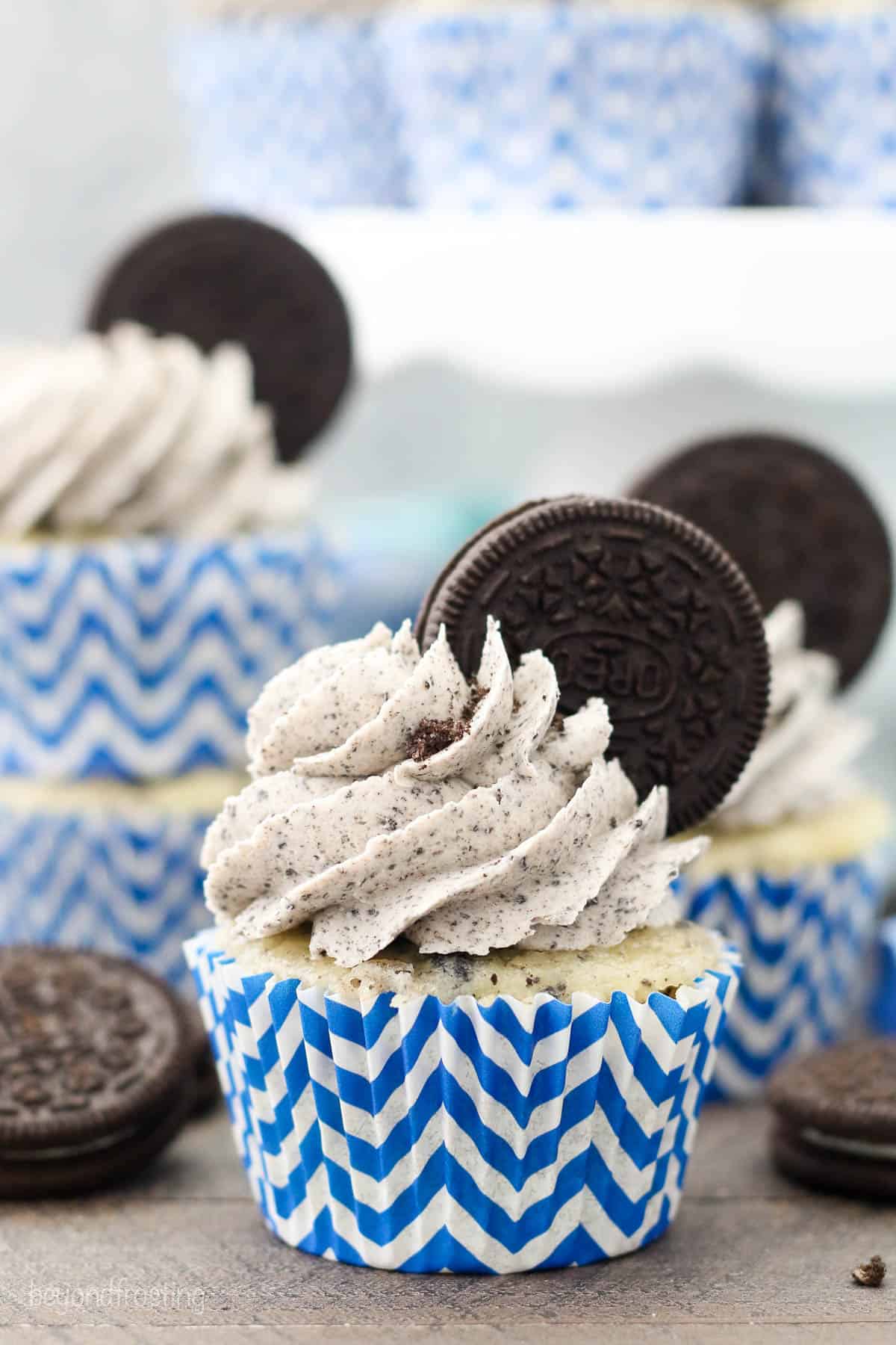 Frosted cookies and cream Oreo cupcakes in white and blue cupcake liners, garnished with whole Oreo cookies.