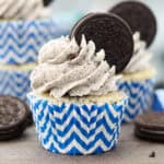 A frosted cookies and cream Oreo cupcake in a white and blue cupcake liner garnished with an Oreo cookie.