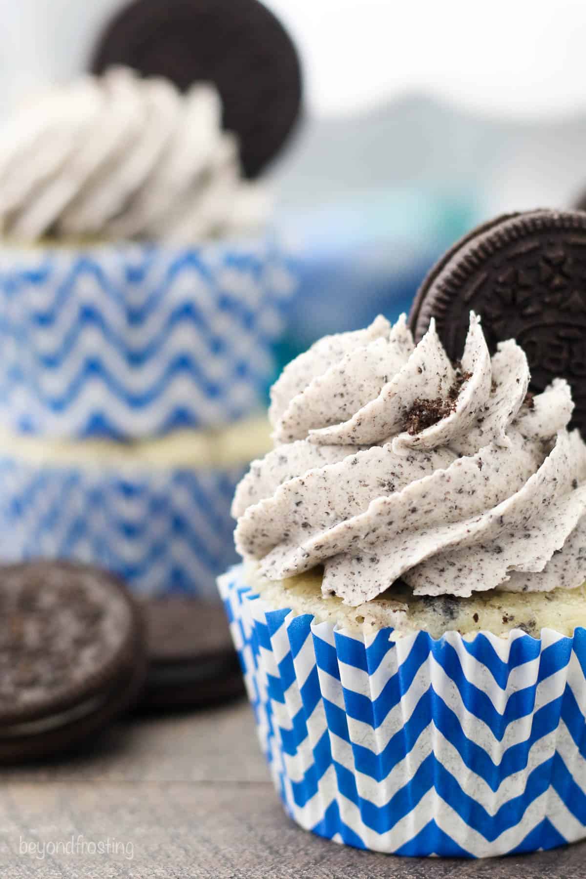 Close up of a frosted cookies and cream Oreo cupcake in a white and blue cupcake liner garnished with an Oreo cookie, with a second decorated cupcake in the background.