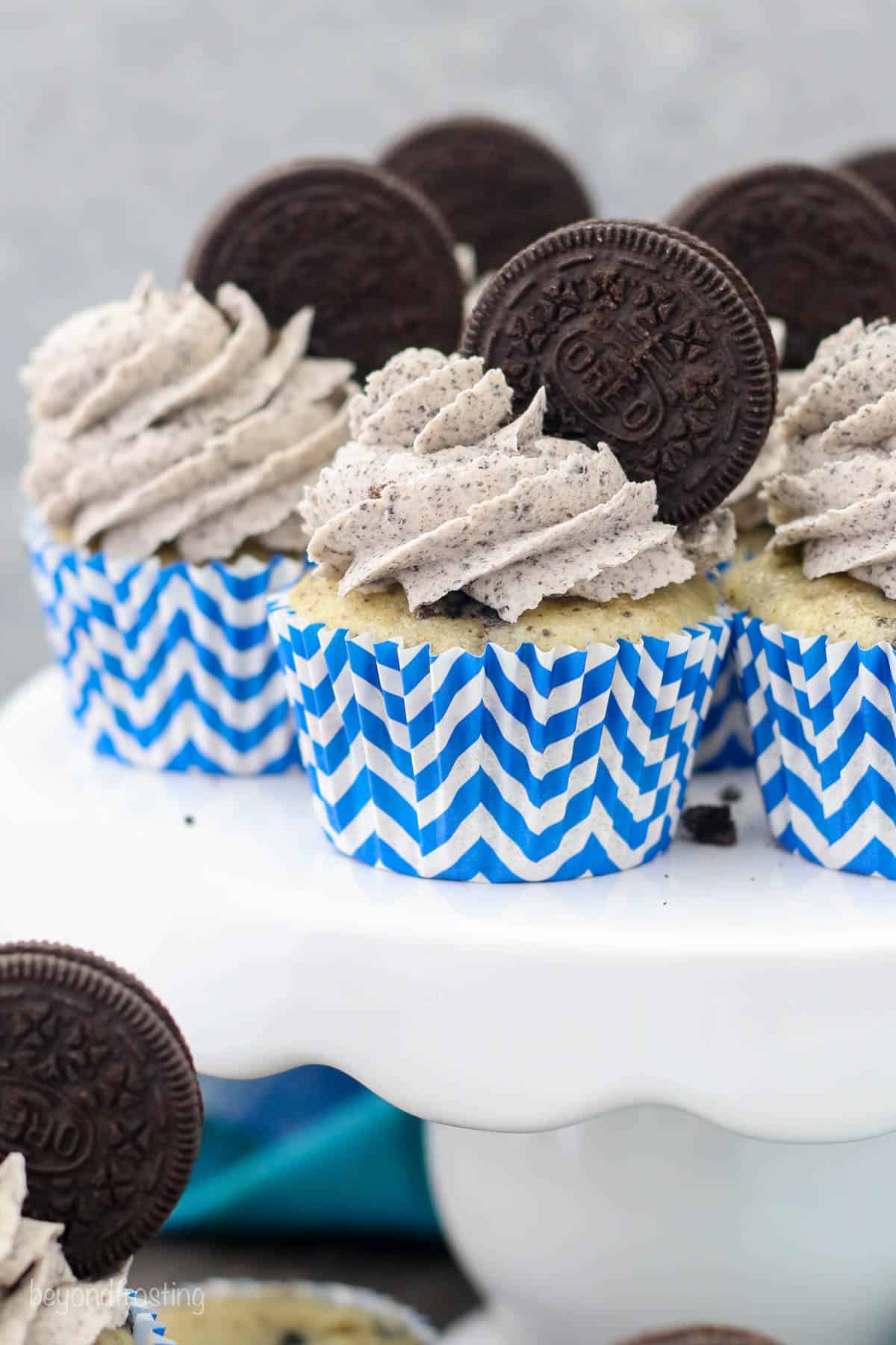 Frosted cookies and cream Oreo cupcakes in white and blue cupcake liners on a white cake stand.