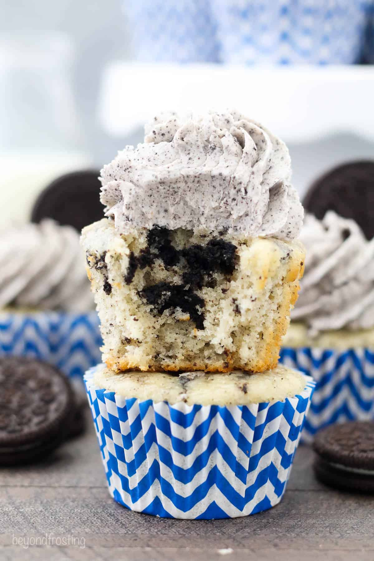 A frosted Oreo cupcake with a bite missing stacked on an unfrosted cupcake, with more decorated cupcakes in the background.