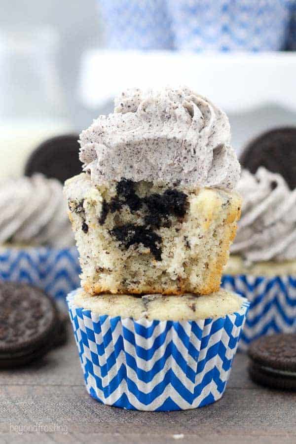 An Oreo cupcake sitting on top of another cupcake. The cupcake has a bite taken out of it, revealing the chunks of Oreo inside the vanilla cupcake. It has an Oreo buttercream