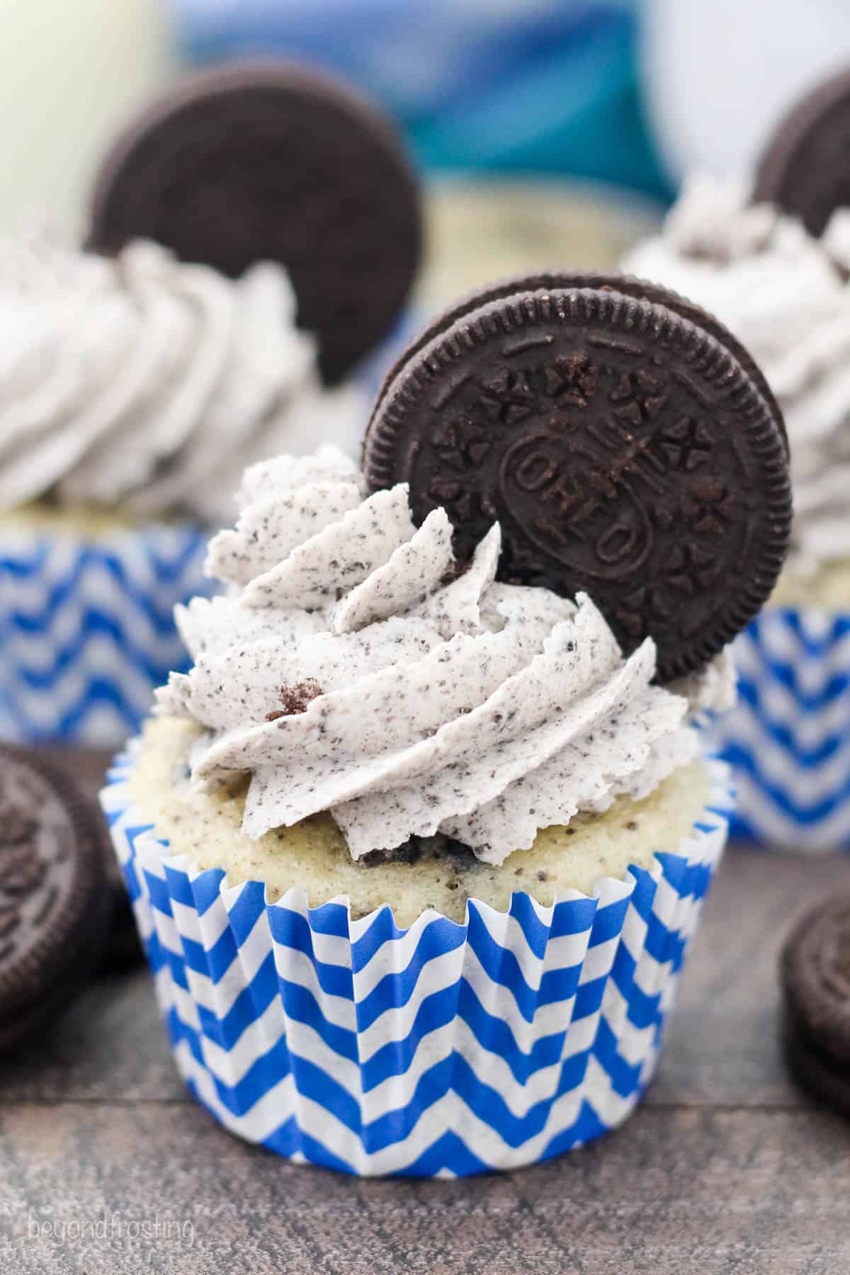 A frosted cookies and cream Oreo cupcake in a white and blue cupcake liner garnished with an Oreo cookie.