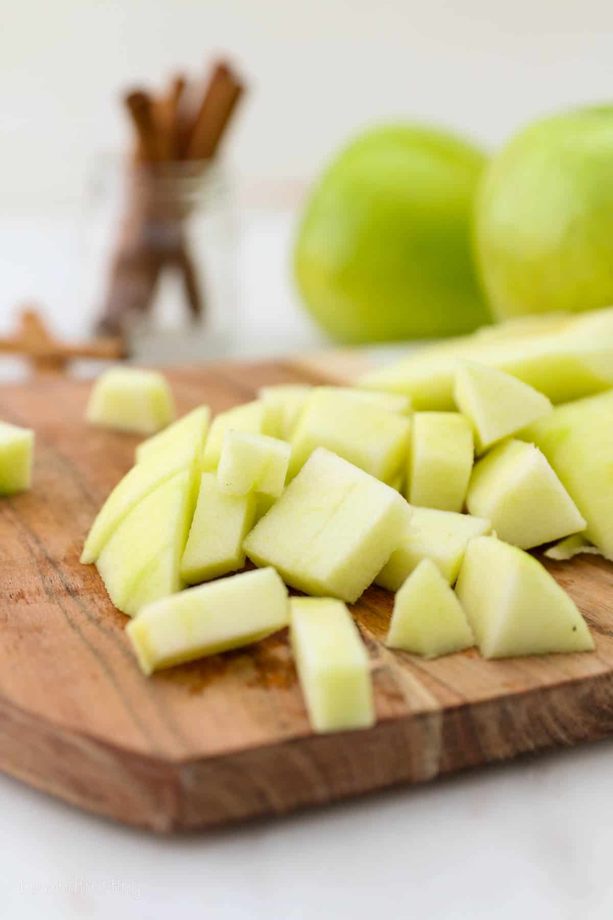 Diced Granny Smith apples on a wooden cutting board, with whole apples and cinnamon sticks in the background.