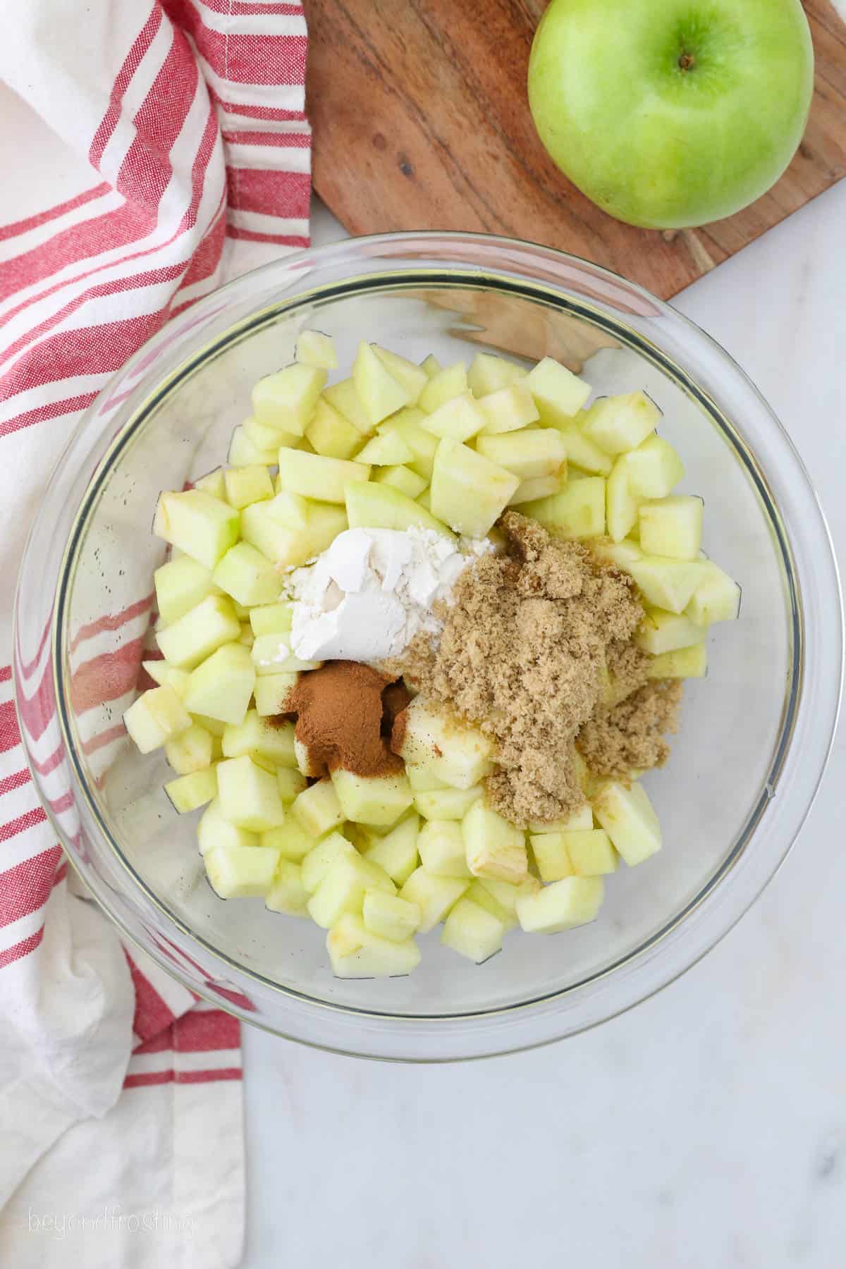 Flour and spices added to a glass bowl with diced apples for apple pie filling.