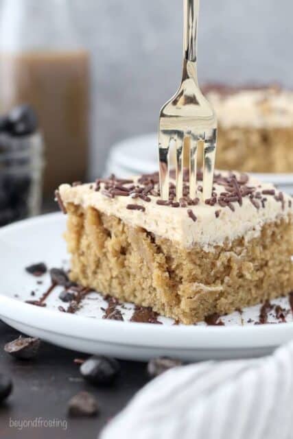 A fork sinking into a slice of mocha cake on a white plate drizzled with chocolate and crushed espresso beans.