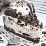 A big slice of No-Bake Oreo Cheesecake on a round white plate drizzled with chocolate and crushed Oreos with a silver fork on the plate.