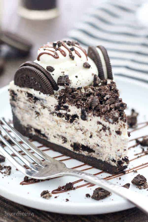 A big slice of No-Bake Oreo Cheesecake on a round white plate drizzled with chocolate and crushed Oreos with a silver fork on the plate.