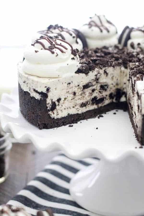 A large Oreo Cheesecake on a white cake plate with a scalloped edge. The cheesecake is garnished with large piles of whipped cream.