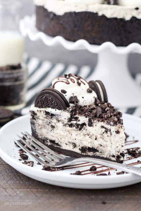 A big slice of No-Bake Oreo Cheesecake on a round white plate drizzled with chocolate and crushed Oreos with a silver fork on the plate.