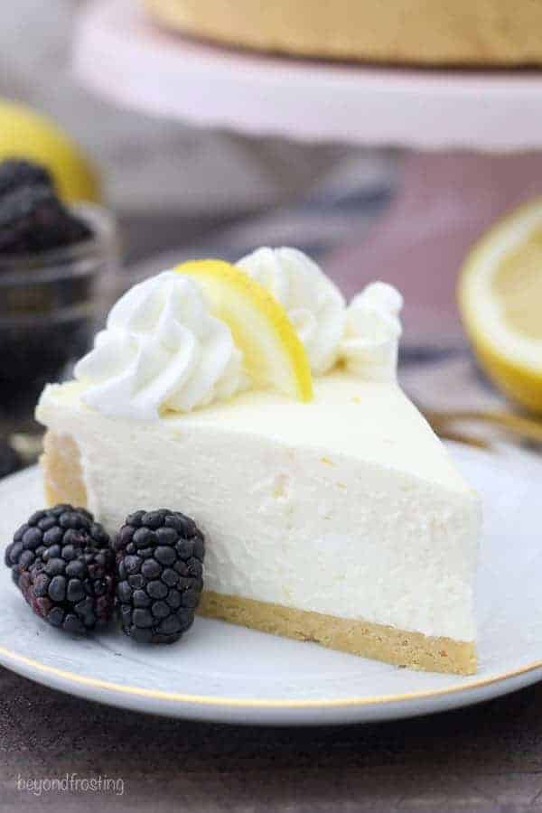 A close up shot of a slide of no-bake lemon cheesecake with two black berries. It's topped with whipped cream and a slice of lemon.