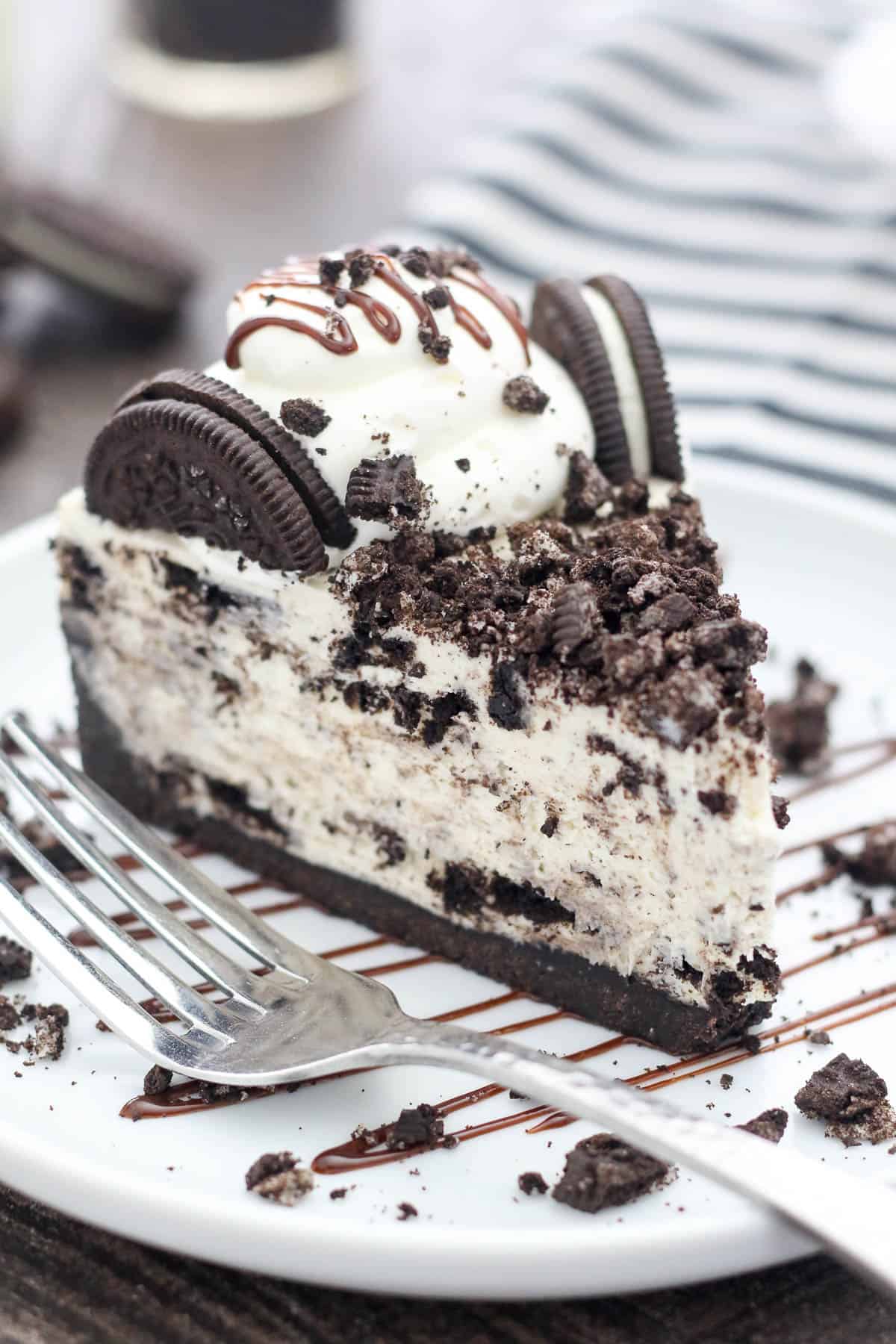 A slice of no-bake Oreo cheesecake on a white plate, topped with Oreos and drizzled with fudge sauce, next to a fork.