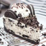 A slice of no-bake Oreo cheesecake on a white plate, topped with Oreos and drizzled with fudge sauce, next to a fork.