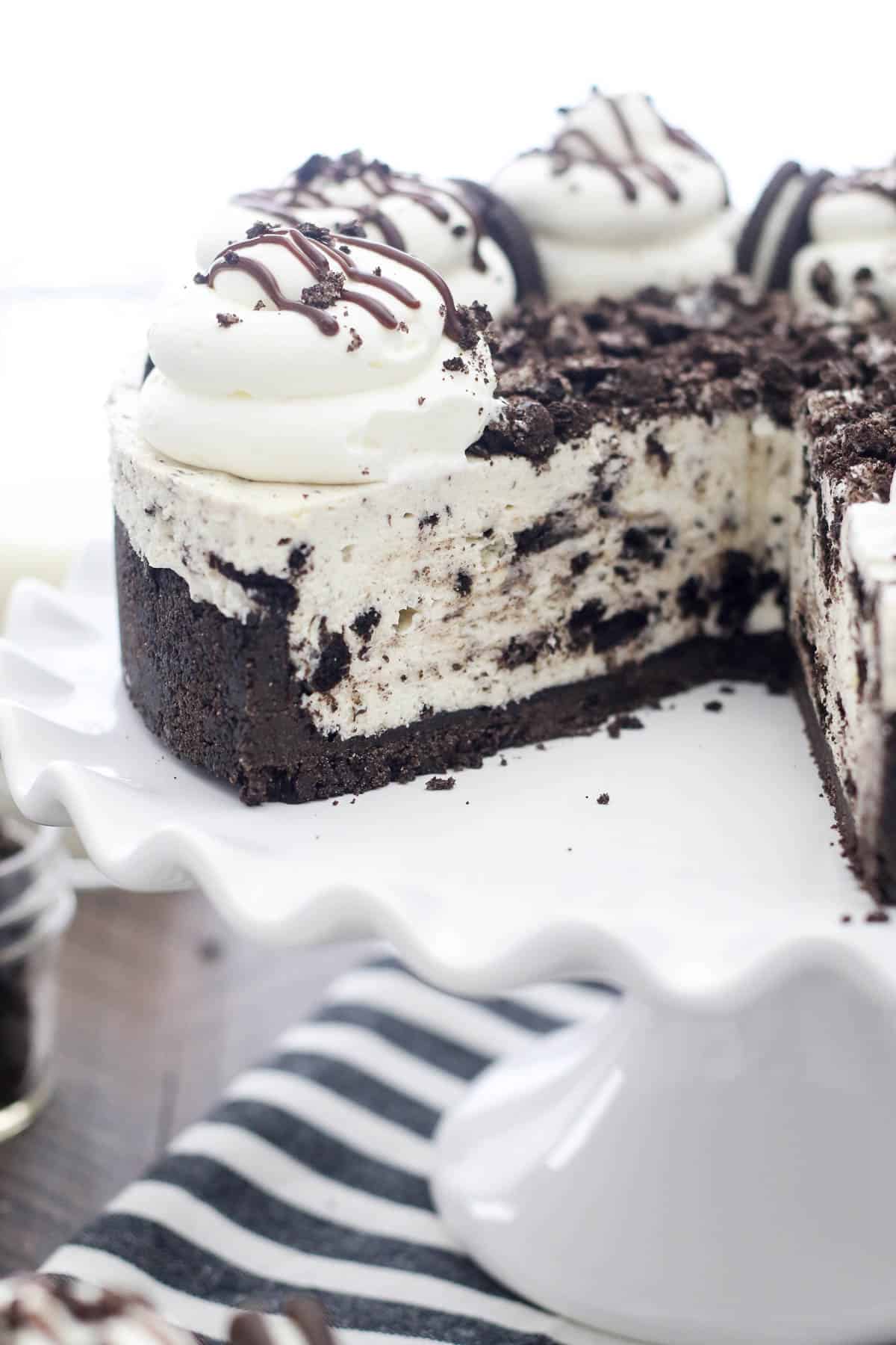 No-bake Oreo cheesecake on a white cake stand, topped with whipped cream swirls, Oreo cookies, and fudge sauce, with a large slice missing.